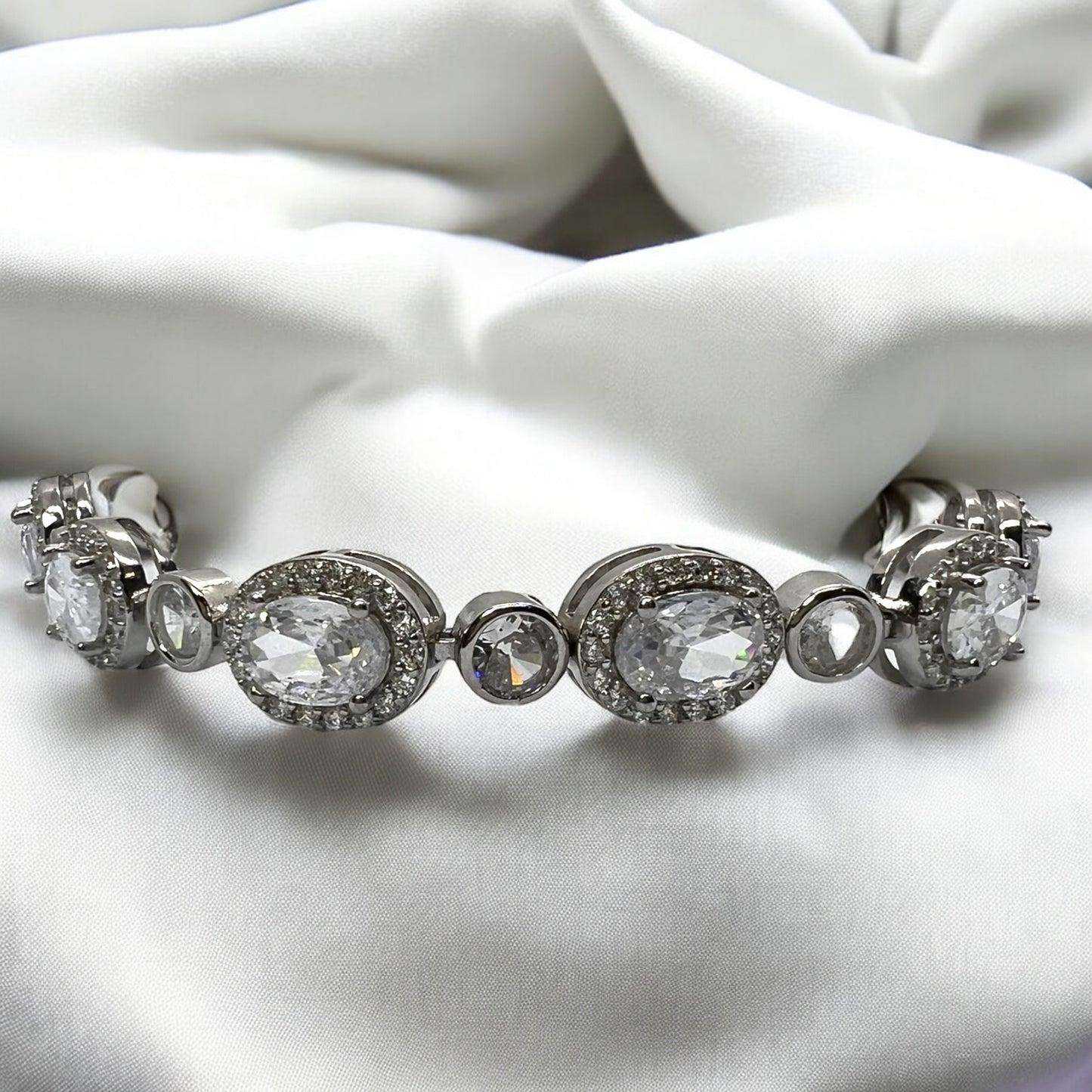 The Velouria Bracelet by Zevaayra, featuring large oval and round simulated diamonds, is elegantly displayed on white satin—an ideal bridal accessory.