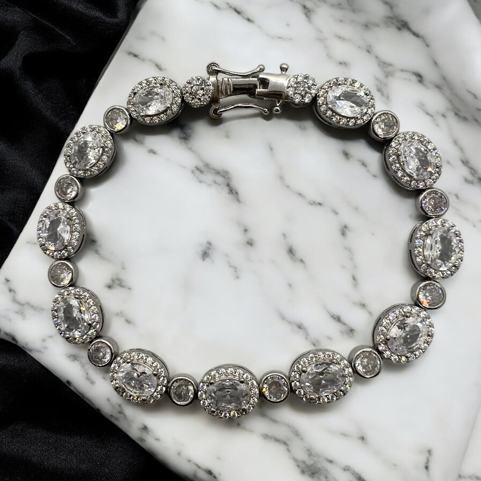 The Velouria Bracelet by Zevaayra features oval and round simulated diamonds set in silver, displayed on white marble with a black satin cloth—an elegant choice for bridal wear.