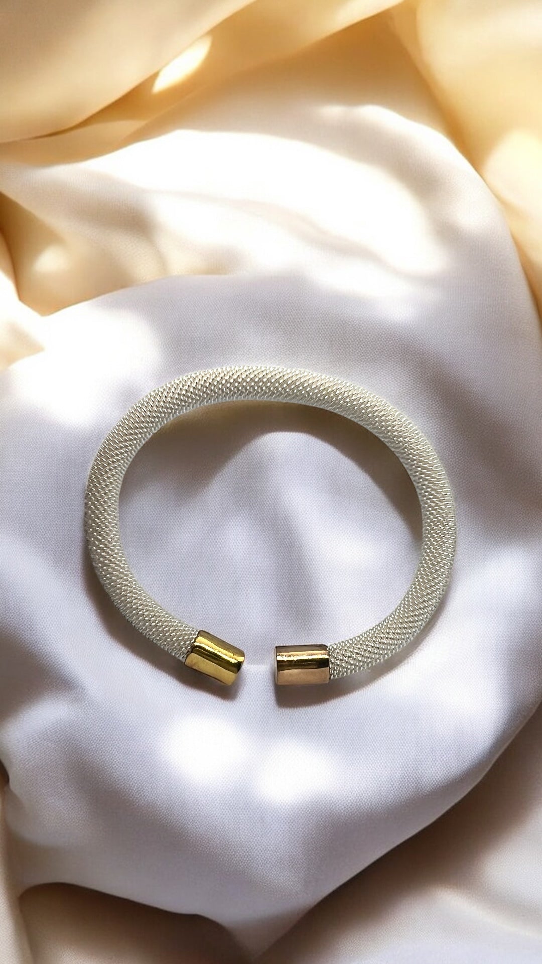 The Zevaayra Luna Mesh Cuff, featuring a textured, tarnish-resistant design with gold-tone end caps, rests in a loose circle atop soft white and cream fabric under gentle lighting.