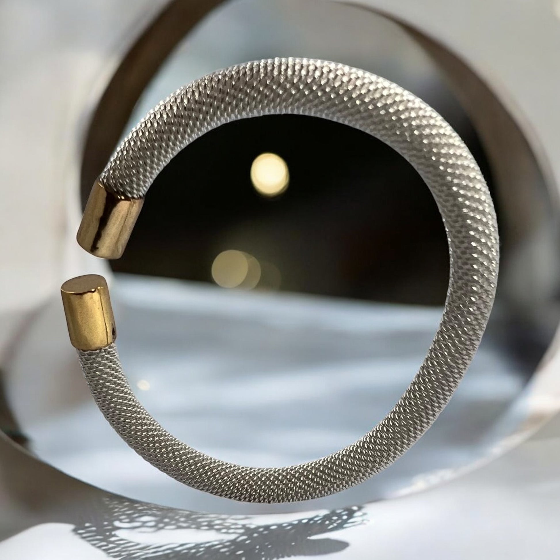 The Zevaayra Luna Mesh Cuff, a tarnish-resistant silver mesh bracelet with gold end caps, is showcased before a circular mirror, creating intricate shadows and reflections on a light surface.