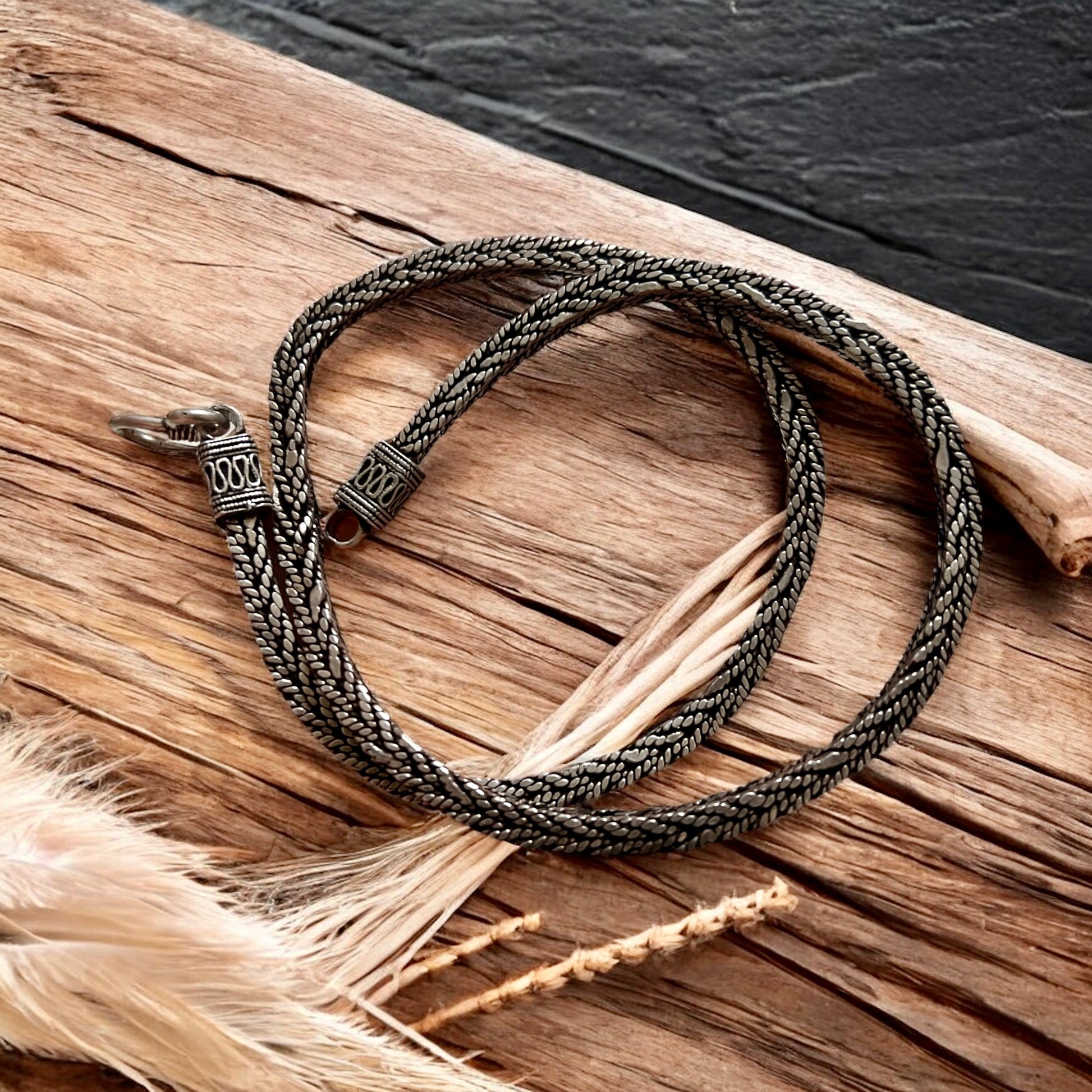The Viraat Chain by Zevaayra, a handcrafted Indian silver herringbone necklace with a decorative clasp, is coiled on rustic wood beside dried grasses against a dark textured background.