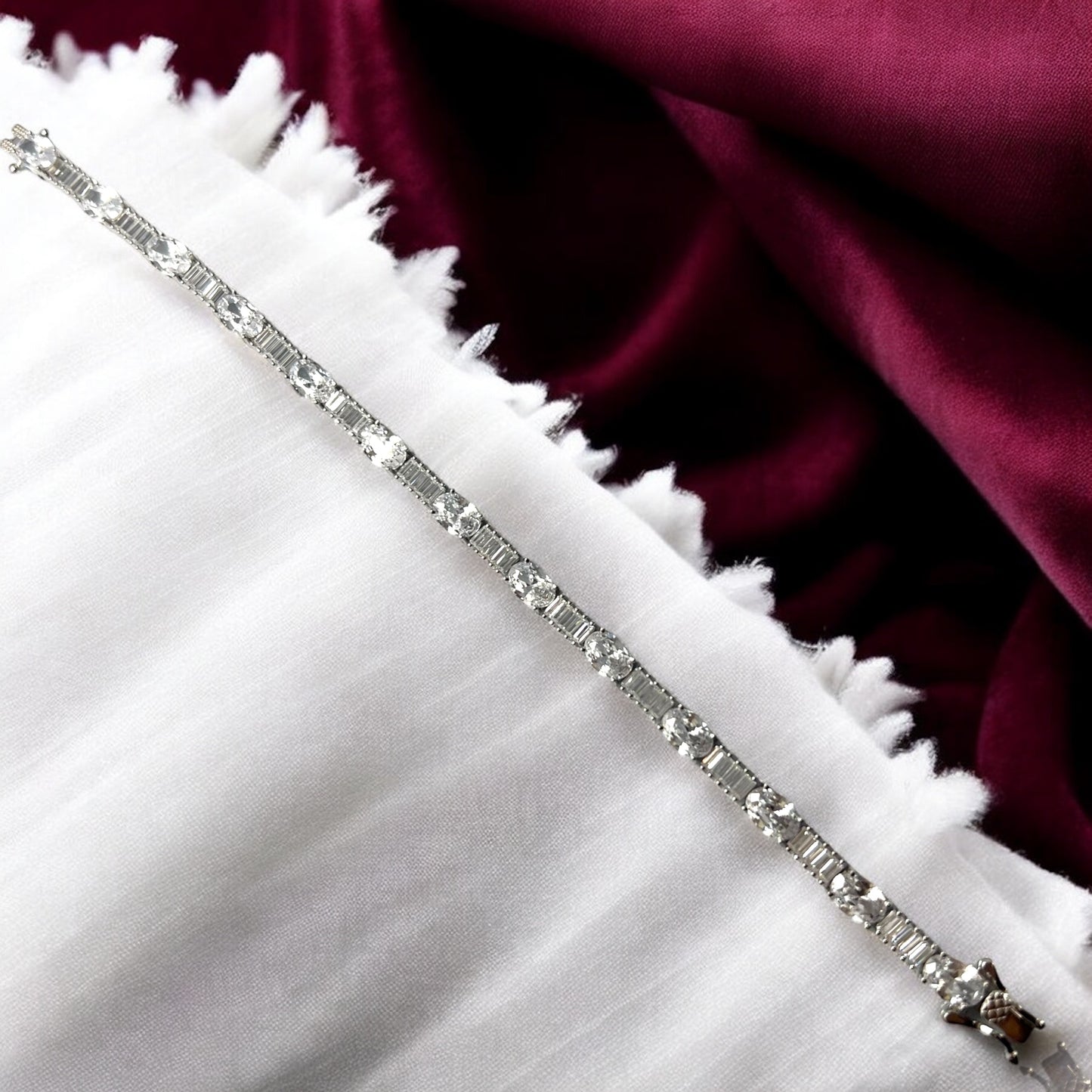 The Zevaayra Victoria Royale Bracelet, featuring oval-cut stones, is displayed on white fringed fabric with a deep burgundy satin background.