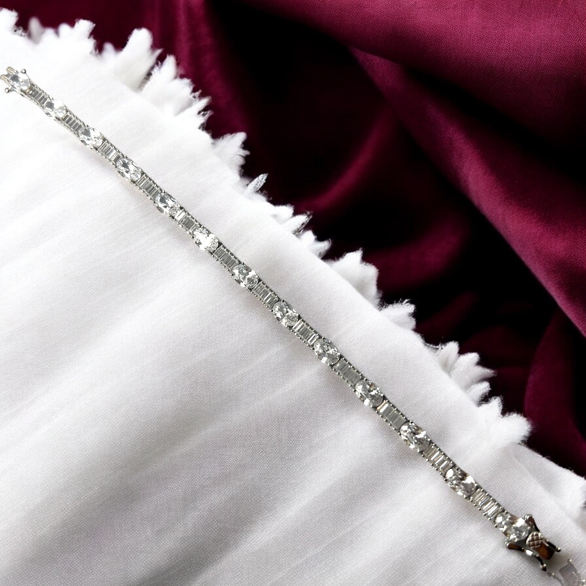 The Zevaayra Victoria Royale Bracelet, featuring oval-cut stones, is displayed on white fringed fabric with a deep burgundy satin background.