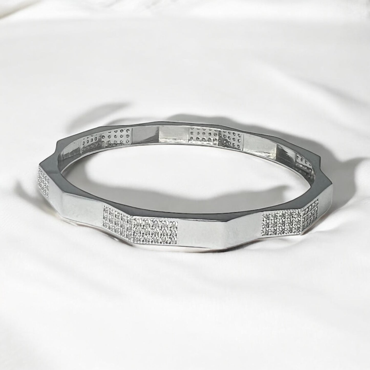 The Zevaayra Zeneer Cuff – Polished Silver Brilliance showcases a geometric, faceted sterling silver design with sparkling cubic zirconia accents, elegantly displayed on soft white fabric.