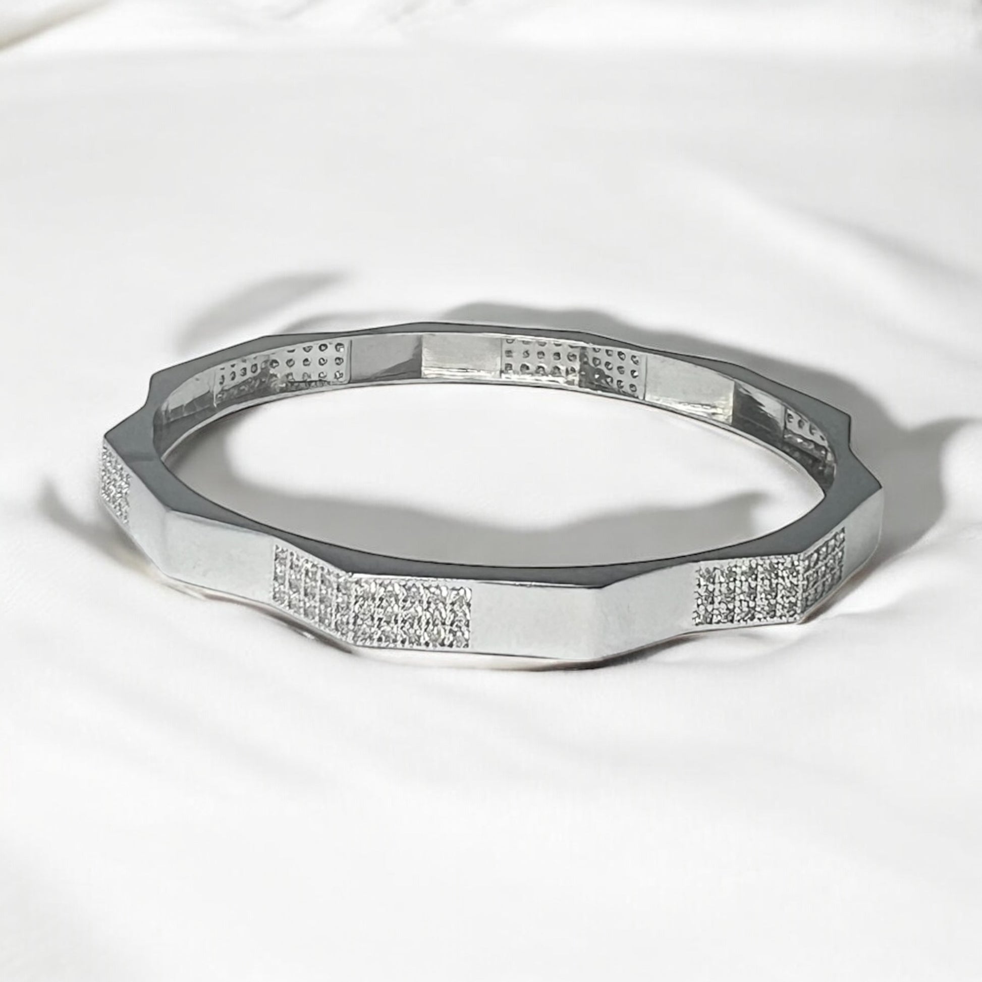 The Zevaayra Zeneer Cuff – Polished Silver Brilliance showcases a geometric, faceted sterling silver design with sparkling cubic zirconia accents, elegantly displayed on soft white fabric.