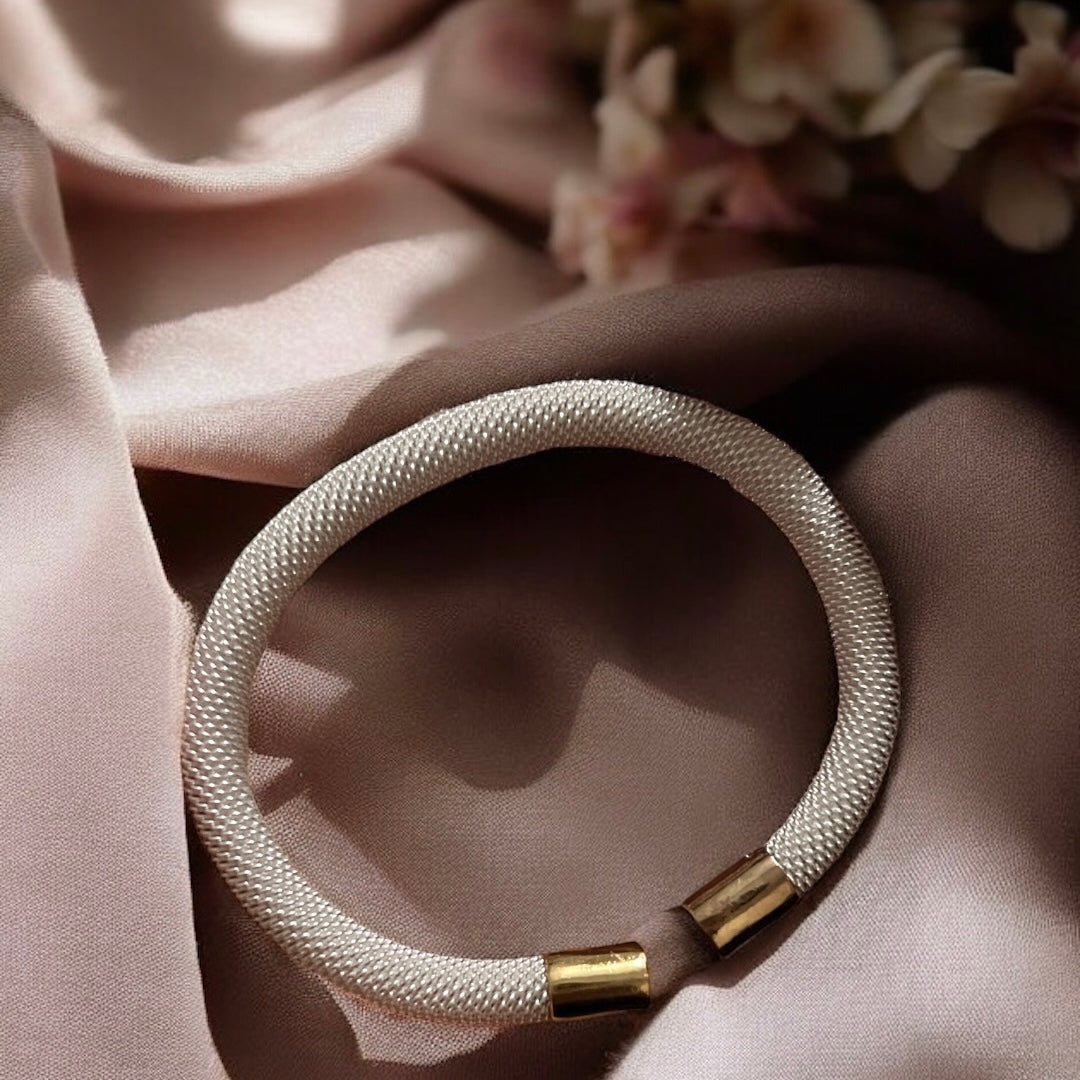 The Zevaayra Luna Mesh Cuff, a white minimalist textured bracelet with gold, tarnish-resistant end caps, rests on blush satin fabric with blurred flowers in the background.