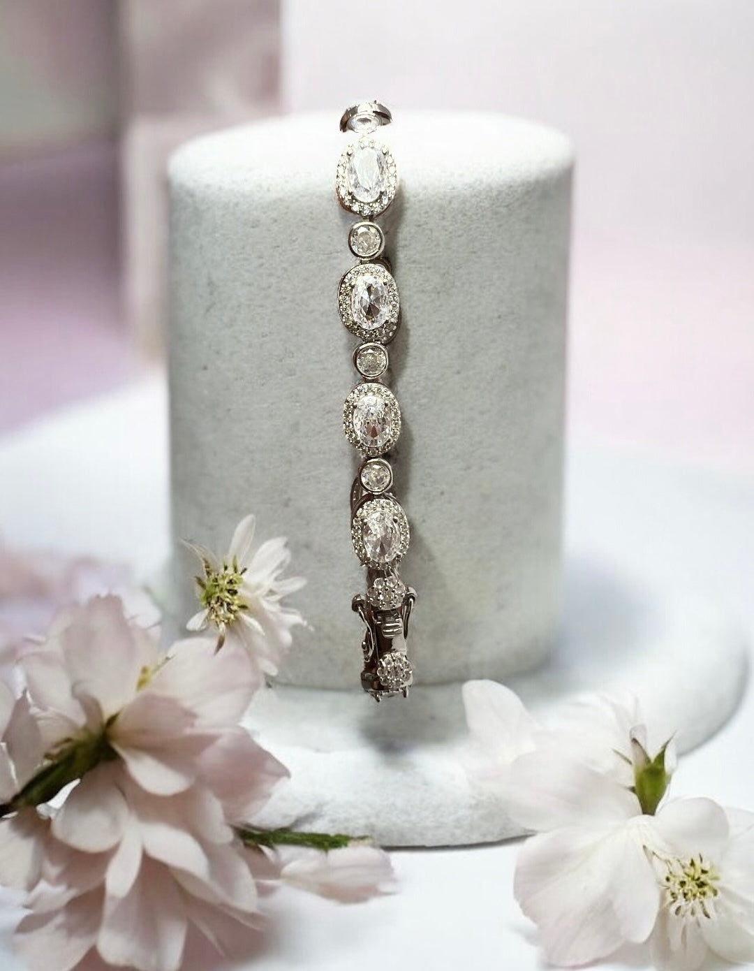 The Velouria Bracelet by Zevaayra, featuring oval and round simulated diamonds, is displayed upright on a white cylindrical stand amid soft pink flowers with a pastel blurred background.