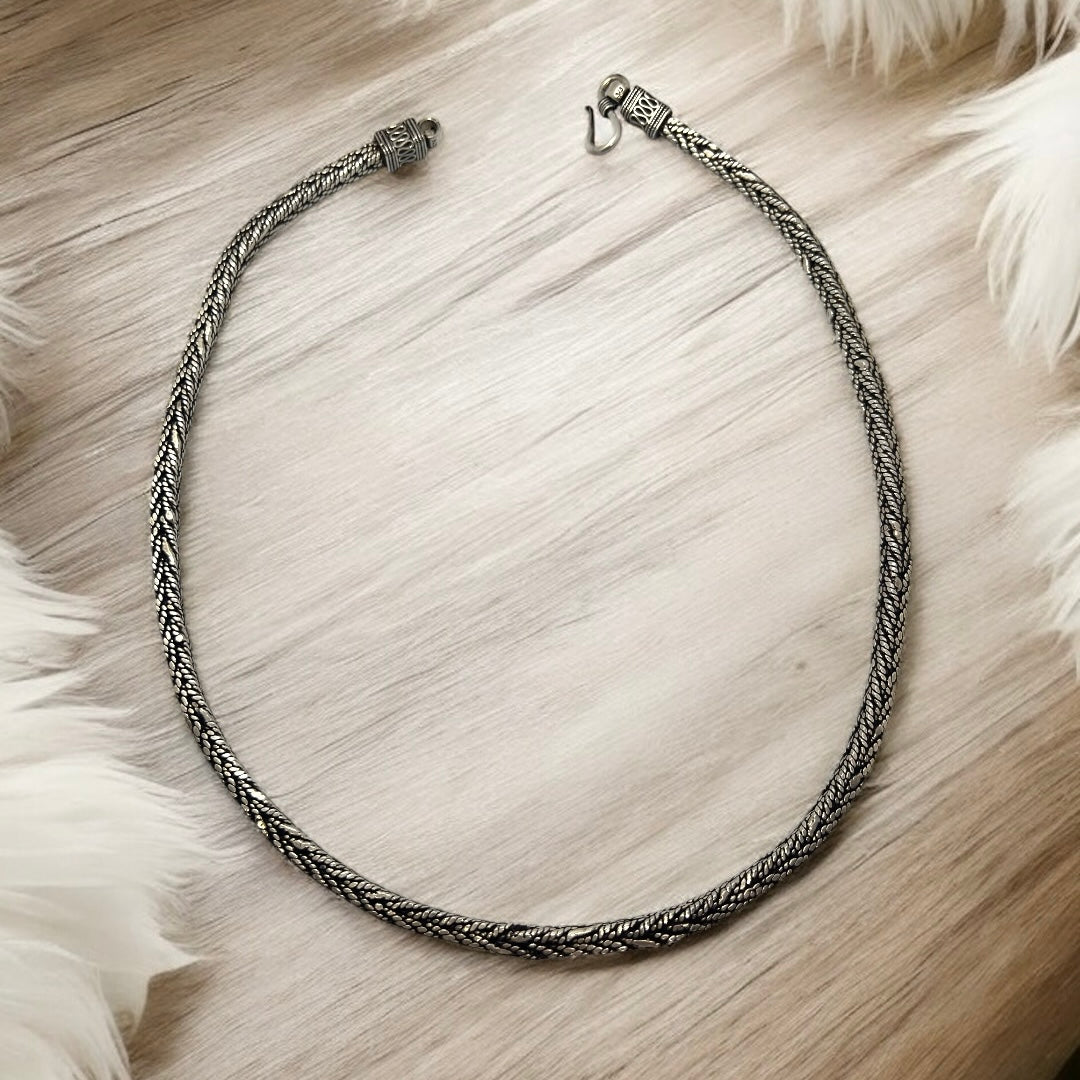 The Zevaayra Viraat Chain, a textured oxidized silver necklace with a detailed pattern, is displayed in a loose oval on a light wooden surface, soft white fur accenting the image’s corners.