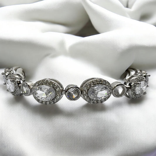 The Velouria Bracelet by Zevaayra, featuring large oval and round simulated diamonds, is elegantly displayed on white satin—an ideal bridal accessory.