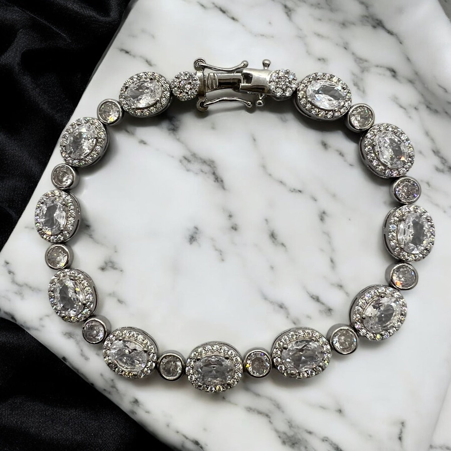 The Velouria Bracelet by Zevaayra features oval and round simulated diamonds set in silver, displayed on white marble with a black satin cloth—an elegant choice for bridal wear.