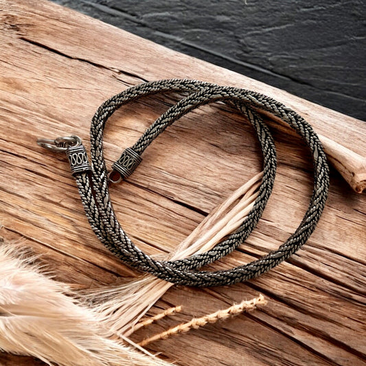 The Viraat Chain by Zevaayra, a handcrafted Indian silver herringbone necklace with a decorative clasp, is coiled on rustic wood beside dried grasses against a dark textured background.