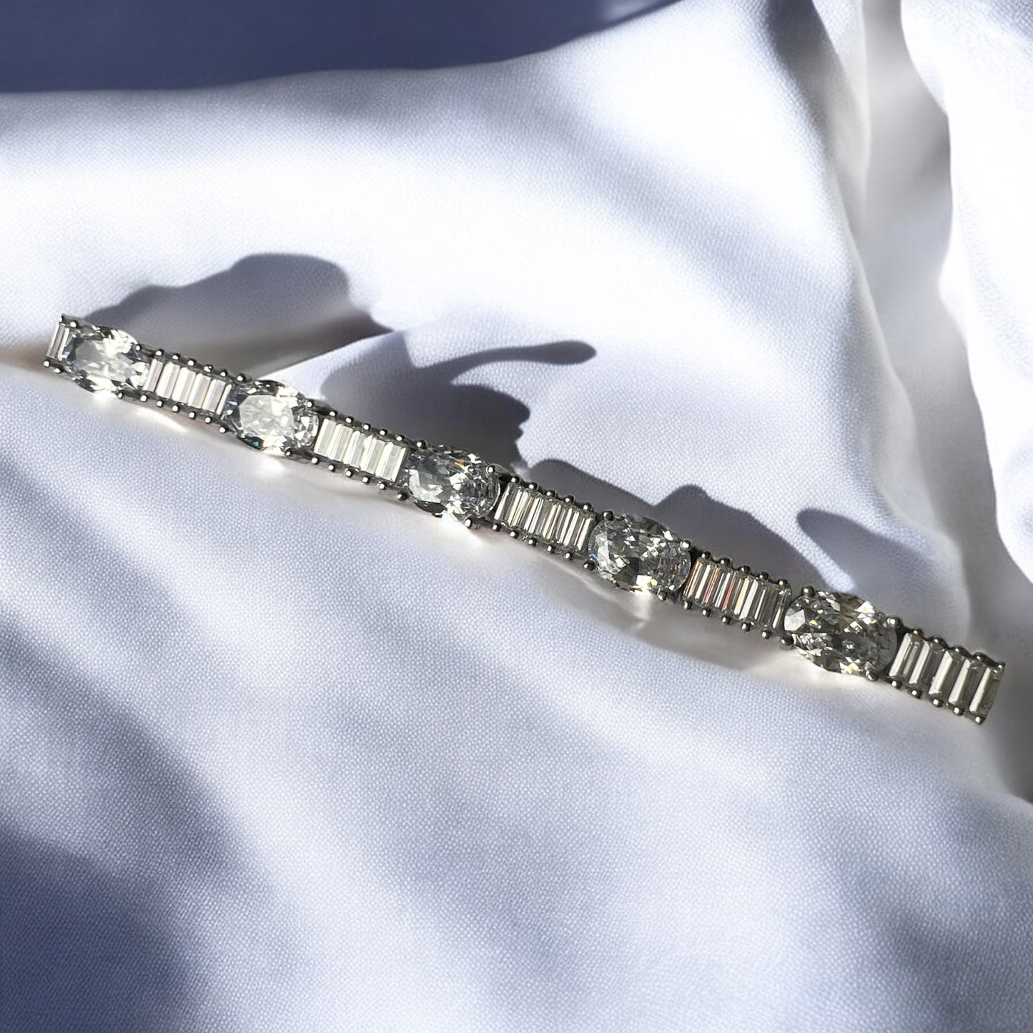 The Zevaayra Victoria Royale Bracelet, featuring alternating round and rectangular clear gemstones in a timeless silver design, rests on softly wrinkled white fabric, gleaming brightly and casting delicate shadows.
