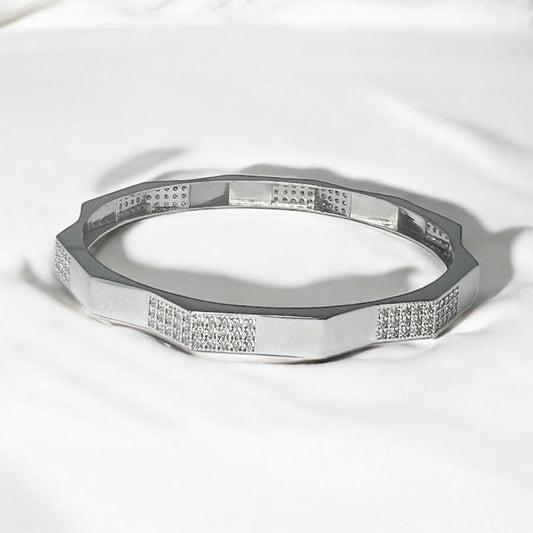 The Zevaayra Zeneer Cuff – Polished Silver Brilliance showcases a geometric, faceted sterling silver design with sparkling cubic zirconia accents, elegantly displayed on soft white fabric.